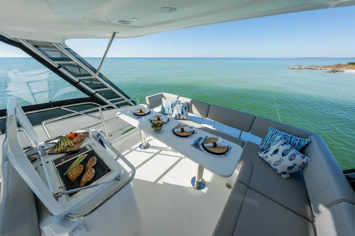 Luxury yacht deck with a grill, dining table set for four, and scenic ocean views under a clear sky.